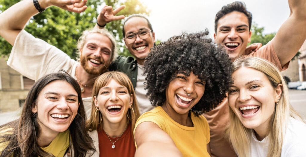 Grupo de jovens multiculturais sorrindo ao ar livre, tirando uma selfie enquanto caminham na cidade. Representação de conexão e bem-estar social, fundamentais para a regulação do estresse.